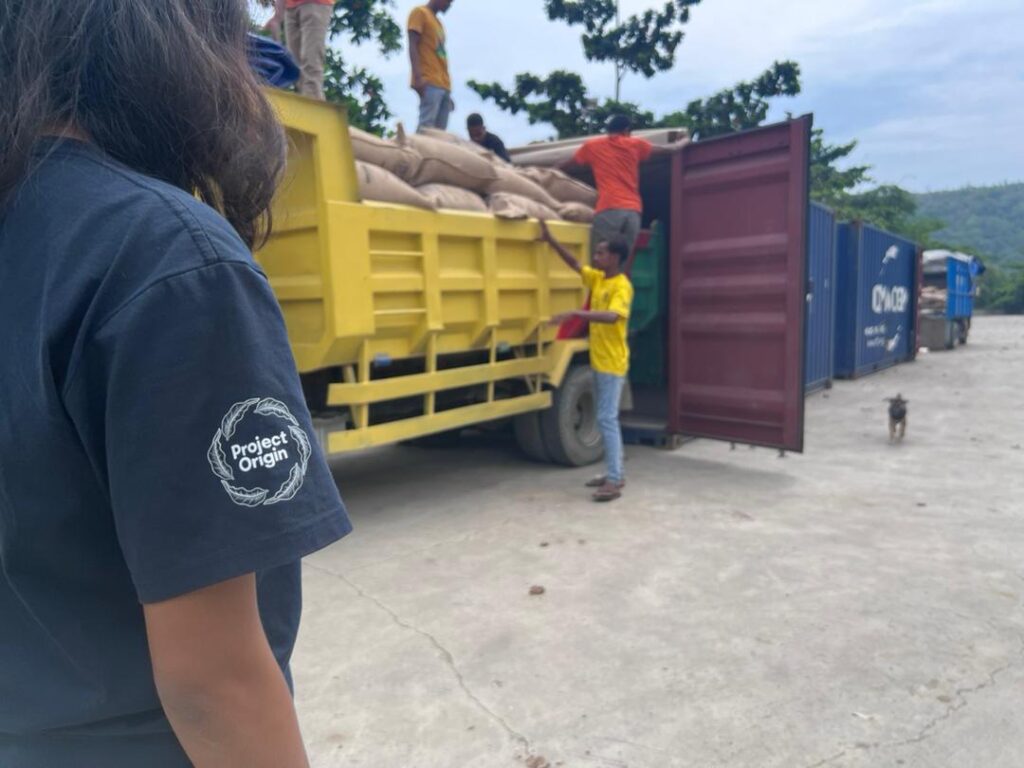 Yellow truck filled with coffee jute sacks backing into a shipping container with a woman in a project origin shirt overseeing progress